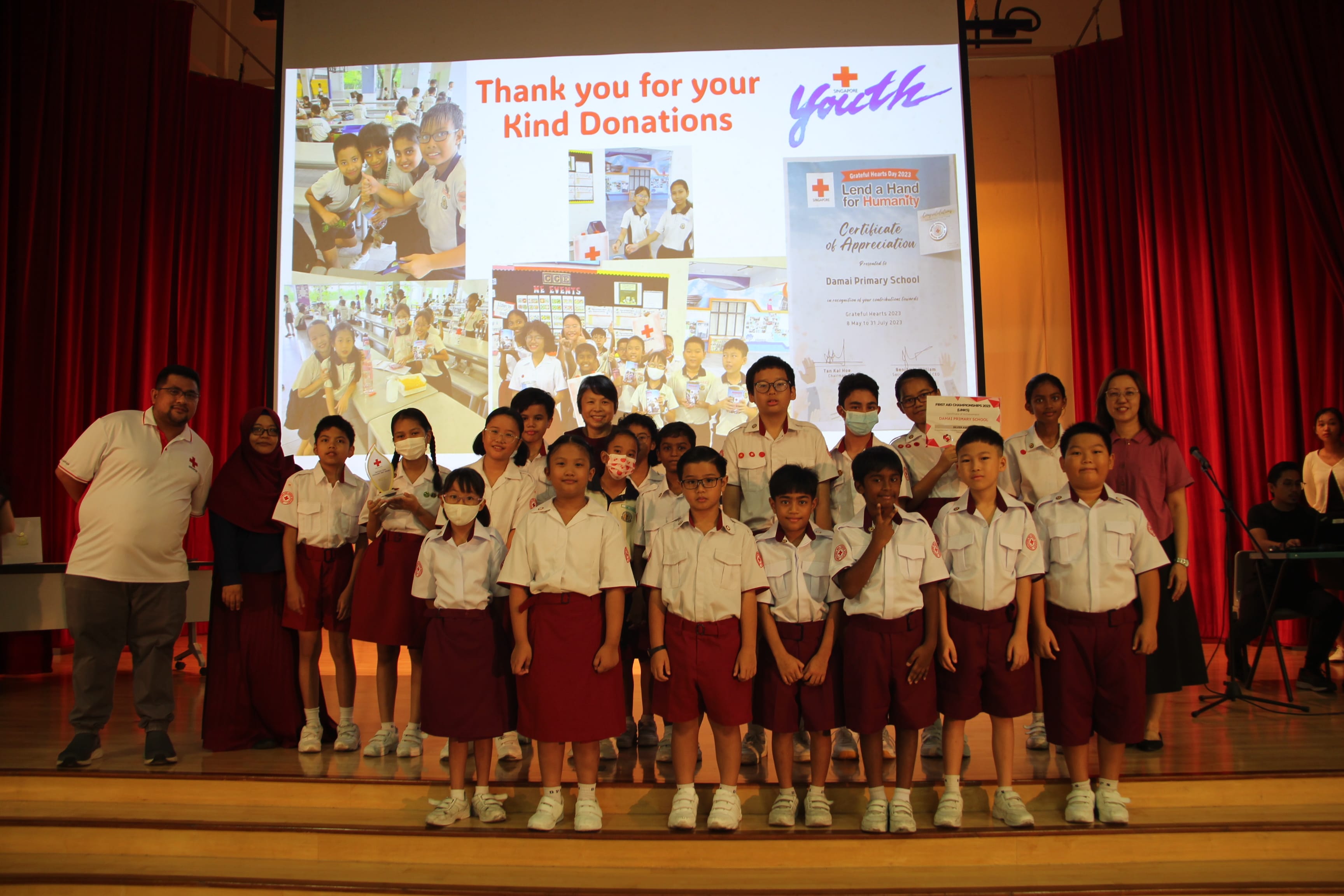Students in Red Cross uniforms stand on a stage in front of a projection reading "Thank you for your Kind Donations."